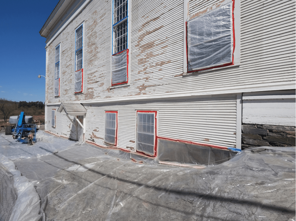A building undergoing lead paint removal, with protective plastic sheeting covering the ground and windows sealed with red tape for safety. A lift and abatement equipment are visible in the background.