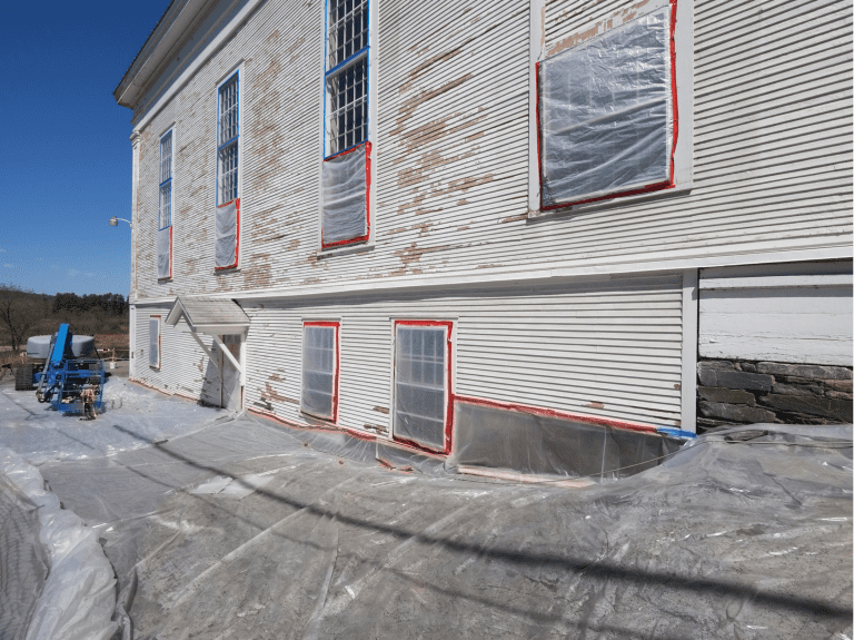 A building undergoing lead paint removal, with protective plastic sheeting covering the ground and windows sealed with red tape for safety. A lift and abatement equipment are visible in the background.